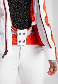 Gros plan sur une personne portant un pantalon de ski blanc et une veste de ski blanche et orange ouverte, dévoilant une couche intérieure orange et une chemise noire à motifs.