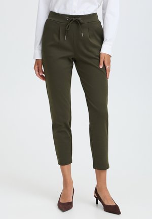 Femme portant un pantalon olive longueur cheville avec taille à cordon, chemise blanche à manches longues, et talons pointus marron foncé, debout.