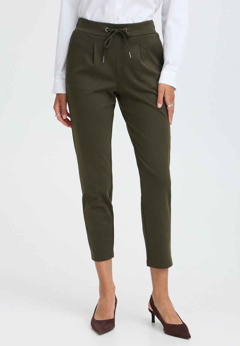 Femme portant un pantalon olive longueur cheville avec taille à cordon, chemise blanche à manches longues, et talons pointus marron foncé, debout.