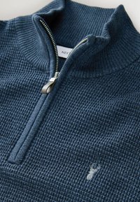 Granatowy sweter zapinany na zamek z teksturowanym wzorem, wyposażony w wysoki kołnierz i srebrny zamek błyskawiczny, ozdobiony małym haftowanym logo.