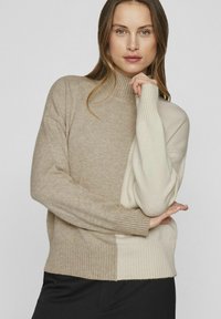 Gebreide trui in beige en crème, met een hoge halslijn, geribde manchetten en onderzoom. Zachte textuur met contrasterende kleurvlakken.