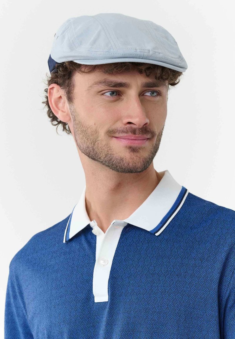 Joven con barba clara que lleva una gorra plana azul claro y una camiseta polo texturizada azul con cuello blanco y tapeta de botones.