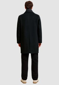 Schwarzer Wollmantel mit strukturiertem Design und hohem Kragen, ausgestattet mit einer sauberen Rückennaht, kombiniert mit schwarzen Hosen und beigen Schuhen.