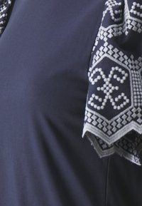 Mörkblått tyg med ett vitt broderat geometriskt och blommönster i hörnet av ett plagg eller textil.