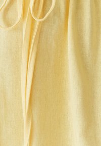 Šviesiai geltona medžiaga su minkštu, tekstūruotu paviršiumi. Matomas raištelis rodo, kad dizainas yra surinktas arba reguliuojamas.