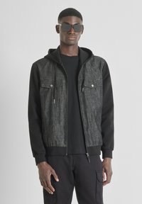 Chaqueta negra con cremallera y parte superior con textura de mezclilla, dos bolsillos frontales y capucha negra. Lleva puesta una camiseta negra y está combinada con pantalones oscuros.
