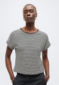 Γυναίκα με μαύρο-λευκό ριγέ κοντομάνικο τοπ και χρυσούς κρίκους σκουλαρίκια, στέκεται με τα χέρια στις τσέπες σκούρων παντελονιών μπροστά σε απλό φόντο.