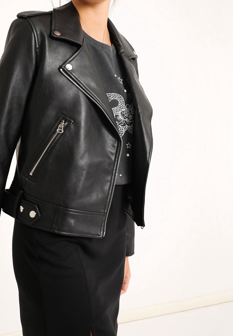 Giacca da motociclista in pelle nera con dettagli in argento, tasche con cerniera e bottoni automatici, indossata sopra una maglietta grafica scura e una gonna nera.