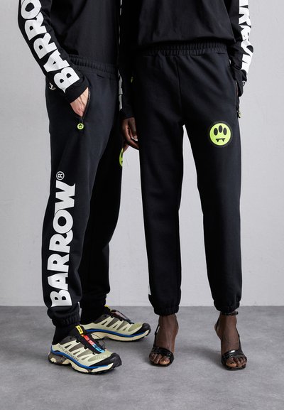Pantaloni sportivi neri con la scritta "BARROW" in bianco, caratterizzati da accenti in verde neon. Una persona indossa sandals neri, mentre l'altra è in scarpe da ginnastica multicolori.