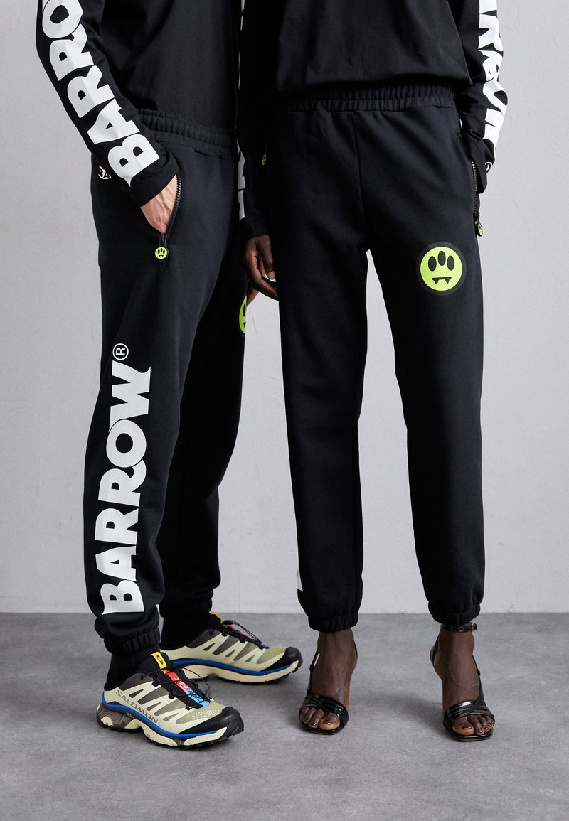Black sweatpants with "BARROW" printed in white, featuring neon green accents. One person wears black sandals, the other is in multi-colored athletic shoes.