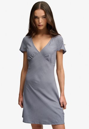 Młoda kobieta z długimi brązowymi włosami, ubrana w szarą sukienkę z krótkim rękawem i dekoltem w serek, z małymi białymi kropkami, stoi na jednolitym białym tle.