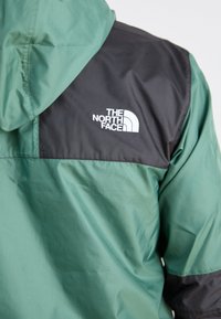 Grüne und schwarze Jacke mit Kapuze. Hergestellt aus glattem Stoff. Mit einem weißen Logo auf der linken Schulter. Kontrastierendes Farbschema.