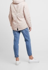 Ljusrosa vadderad jacka med huva, med en delad bakdesign, bärs över något avsmalnande blå jeans och vita sneakers.