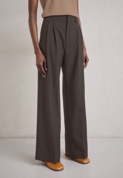 Pantaloni a vita alta e gamba larga in tessuto marrone scuro con pieghe frontali. Abbinati a un top beige e scarpe con tacco marroni. Tessuto liscio.