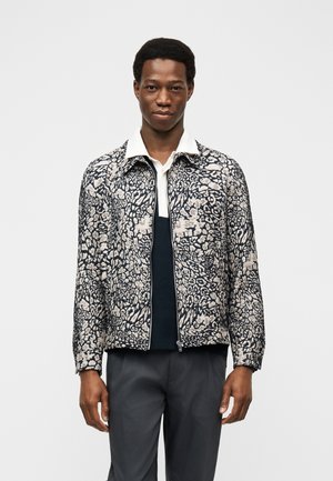 Mand iført en sort og hvid blomsterprintet lynlåstrøje over en hvid skjorte med krave og mørke bukser, stående foran en hvid baggrund.