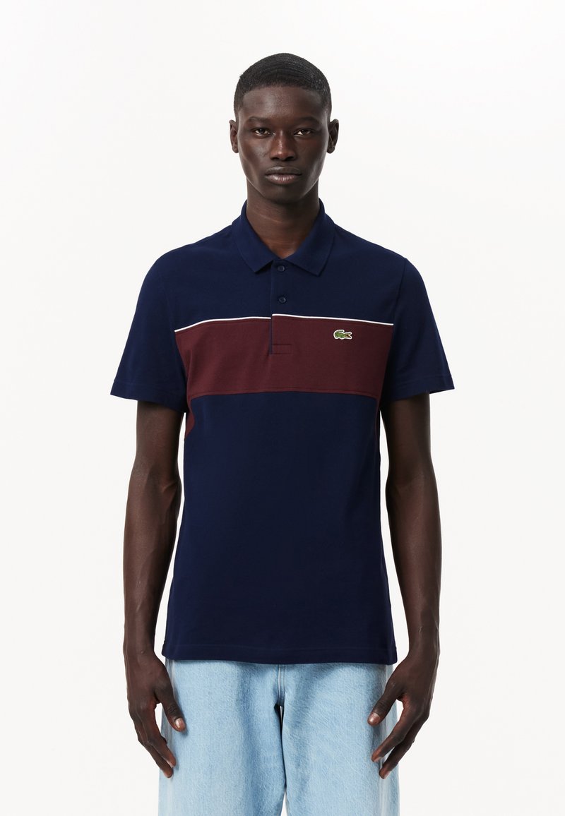 Laivastonsininen polo-paita, jossa on viininpunainen raita ja valkoiset yksityiskohdat. Valmistettu tekstuurisesta puuvillasta, jossa on kaksipainikkeinen kaulus ja vihreä logo.