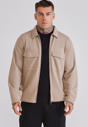 Homme avec des tatouages au cou et aux mains, portant une veste beige zippée par-dessus un t-shirt noir, debout devant un fond gris clair uni.