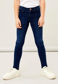 Kék farmer skinny farmernadrág öt zsebes kialakítással, övbújtatókkal és elöl gombos záródással. Fehér sportcipővel viselve.