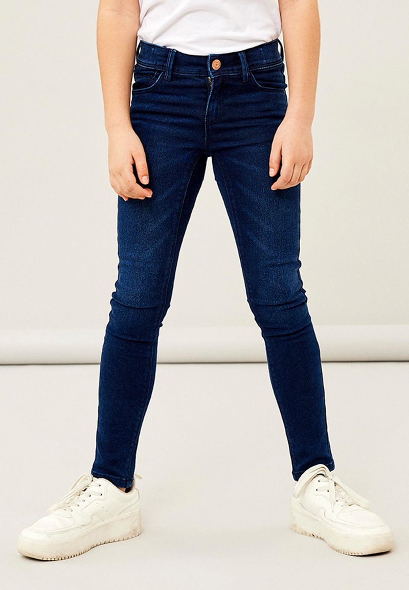 Kék farmer skinny farmernadrág öt zsebes kialakítással, övbújtatókkal és elöl gombos záródással. Fehér sportcipővel viselve.