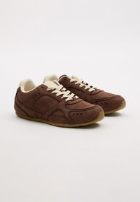 Zapatillas de ante marrón con cordones y acentos en beige. Presentan perforaciones en los lados, suela de goma y un interior acolchado para mayor comodidad.