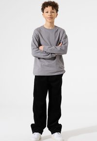 Grijze langemouwen shirt met een ronde hals, gecombineerd met zwarte corduroy broek. Witte sneakers maken de outfit compleet. Model staat met zijn armen gekruist.