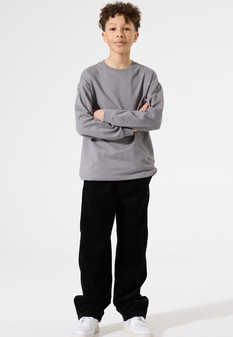 Grijze langemouwen shirt met een ronde hals, gecombineerd met zwarte corduroy broek. Witte sneakers maken de outfit compleet. Model staat met zijn armen gekruist.