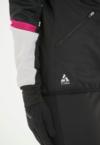 Svart jacka med rosa och vita accenter; har en zippered ficka, WX-TECH-logotyp och åtsittande ärmslut. Handsskarna är också svarta och texturerade.