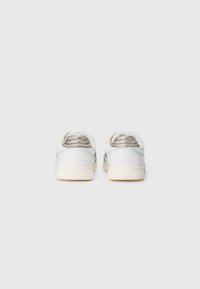 Ett par vita låga sneakers med metallicsilverfärgade vadderade hälkragar visade framifrån på en enkel ljus bakgrund.
