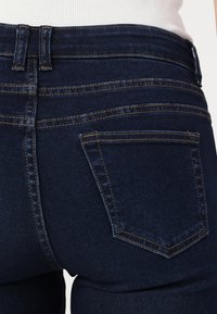 Dark blue denim jeans with a fitted design, featuring gold stitching and a classic back pocket with a pointed flap.