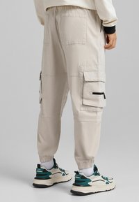 Beige cargo kalhoty s elastickým pasem, několika kapsami včetně stylu cargo a úzkými lemy, ve spojení s černými a tyrkysovými sportovními botami.