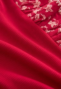Tissu rouge avec une texture côtelée et un motif floral en rouge plus clair. Le design comprend à la fois des sections lisses et froncées.