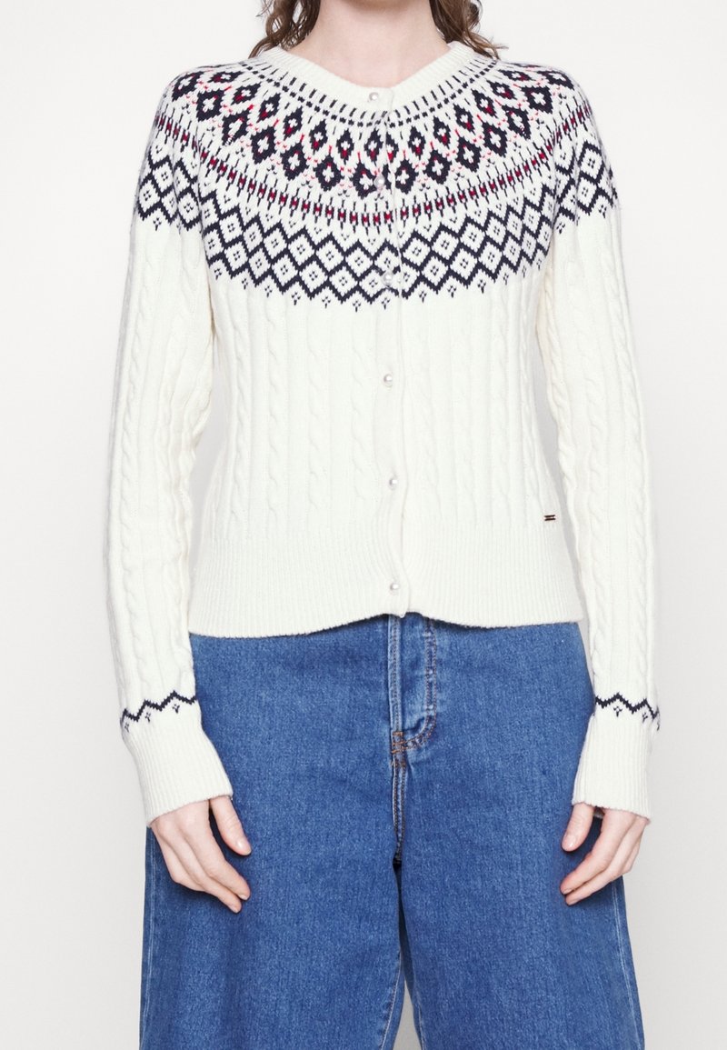 Cardigan blanc en maille torsadée avec motif géométrique noir et rouge sur le yoke, boutonné à l'avant, associé à un jean taille haute bleu.