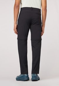 Pantalons noirs extensibles avec des poches latérales zippées ; comprend des jambes inférieures amovibles pour se convertir en shorts. Portés avec des chaussures de sport bleues.