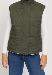 Olivgrüne wattierte Weste mit hohem Kragen, geometrischem Muster, seitlichen Taschen und einem weichen Innenfutter. Getragen über einem weißen Oberteil.