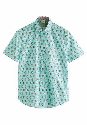 Homme portant une chemise bleu clair à manches courtes avec un motif floral, un short mauve et des sandales marron, debout avec une main dans la poche.