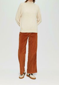 Pull en tricot beige à col montant, assorti d'un pantalon large en velours côtelé rouille. Mis en valeur par des chaussures bicolores avec des lacets noirs.