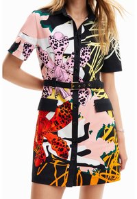 Donna che indossa un vestito abbottonato con cintura, decorato con grandi stampe floreali astratte nei colori rosa, rosso, giallo, nero e bianco, con maniche corte.