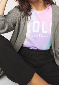 En person som bär en grå stickad cardigan över en pastellfärgad tie-dye Ralph Lauren Polo-tröja och svarta byxor, sitter med ett ben böjt.