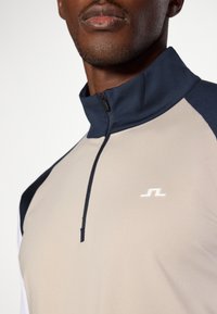 Μπεζ και ναυτικό μπλουζάκι �πόλο με ψηλό γιακά, διαθέτει φερμουάρ και ένα μικρό λευκό λογότυπο στο στήθος. Απαλή ύφανση με αντίθετα χρώματα.