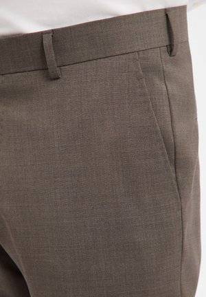 Close-up of brown tailored pants with side pockets and belt loops, worn with a white shirt tucked in.