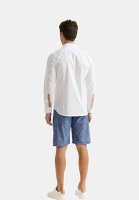 Camicia bianca a maniche lunghe con polsini abbottonati, abbinata a pantaloni corti blu a fantasia e sneakers bianche, vista da dietro.