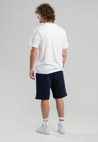 White cotton T-shirt with short sleeves, paired with navy shorts. Model wears white sneakers with green-striped socks, facing away from the camera.