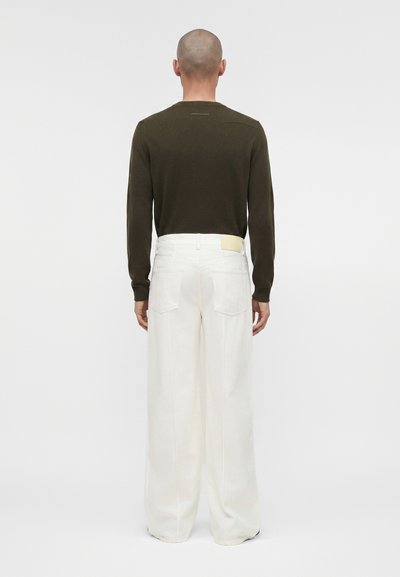 Άτομο με ξυρισμένο κεφάλι στέκεται πλάτη, φοράει σκούρο πράσινο μακρυμάνικο πάνω μέρος και φαρδιά λευκά παντελόνια σε απλό φόντο.