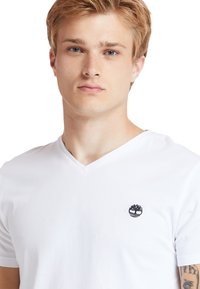 Vit bomull T-shirt med V-ringning och korta ärmar, med en svart logotyp på vänster bröst. Slät textur, figursydd design.