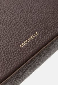 Ruda tekstūruota odinė rankinė su auksiniu "Coccinelle" ženklu paviršiuje. Lygus paviršius ir subtilus grūdų raštas. Stačiakampio formos.