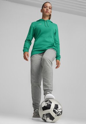 Grüne Kapuzenjacke mit Kängurutasche, kombiniert mit grauen Jogginghosen. Weiße Sportschuhe und ein schwarz-weißer Fußball auf dem Boden.