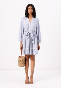 Abito a righe blu e bianche con cintura da annodare, maniche lunghe, orlo arricciato, bottoni frontali e dettagli intricati; abbinato a una borsa in tessuto intrecciato.