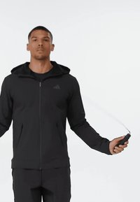 Muž v černé sportovní teplákové soupravě s kapucí skákající přes švihadlo na jednobarevném bílém pozadí.