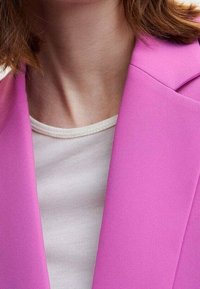 Różowa marynarka o zdefiniowanej konstrukcji i ostrych klapach, nałożona na kremowy, krótki top z rękawami. Widoczna gładka struktura materiału.