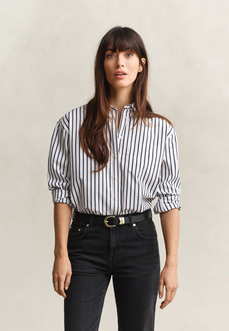Young woman with long brown hair wearing a white shirt with black vertical stripes and black jeans against a plain background.